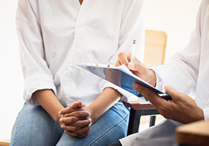 a woman sits with hands clasped on her knee while talking with a medical professional